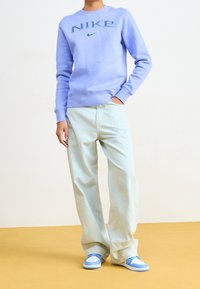 En person som bär en ljuslila Nike-tröja, lösa ljusblå jeans och matchande ljusblå och vita sneakers står på ett gult golv.