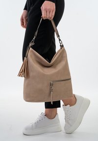 Bolso de hombro de cuero beige con un diseño relajado, bolsillo con cremallera y un detalle de borlas. Combinado con zapatillas blancas y pantalones negros.