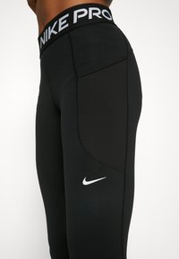 Mustat urheiluhousut, joissa on leveä vyötärönauha, jossa lukee "NIKE PRO" valkoisella. Housut ovat sileää kangasta ja niissä on pieni valkoinen Nike-logo helmassa.