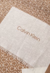 Sciarpa marrone con un motivo a monogramma CK ripetuto, caratterizzata da un bordo bianco con "Calvin Klein" stampato in marrone chiaro. Bordo frangiato.