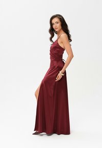 Burgundská saténová večerní toaleta s přiléhavým tělem, tenkými ramínky, bočním rozparkem a nařasenými detaily, s uvázáním vzadu.