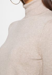 Vaaleanbeige poolopaita pehmeästä neuloskankaasta, jossa on resorikaulus ja sävyyn sopiva omistus. Lähikuva kaula-aukon ja olkapään alueelta.