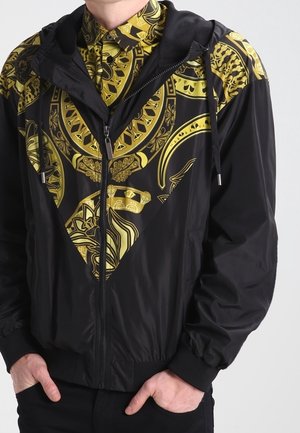 Personne portant une veste noire zippée ornée de motifs dorés élaborés, sur une chemise à boutons assortie avec des motifs dorés et noirs.
