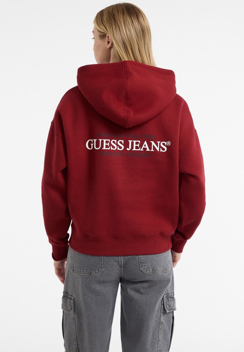 Bordová mikina s veľkým bielym logom "GUESS JEANS" na chrbte. Tkanina je mäkká a obsahuje kapucňu so sťahovacou šnúrkou a rebrované manžety.