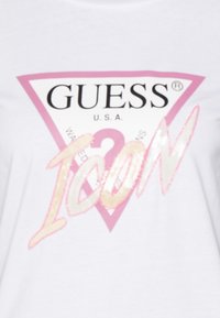 Hvid T-shirt med en trekantet grafik. "GUESS" i fed sort tekst, "U.S.A." nedenunder. Ordet "ICON" i blanke pink og guld bogstaver.