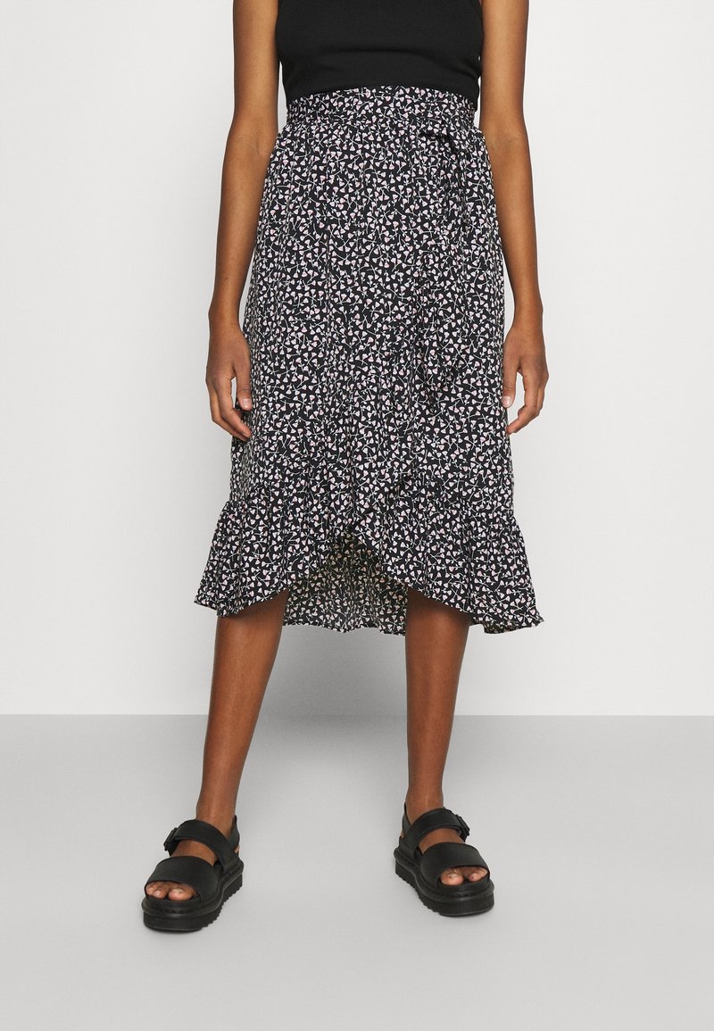 Femme portant une jupe midi noire à fleurs avec un ourlet à volants, un top noir sans manches et des sandales compensées noires, debout devant un fond uni.