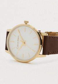 Montre-bracelet dorée avec un cadran blanc texturé, un bracelet en cuir brun et des marqueurs horaires minimalistes. Dispose d'une couronne proéminente.