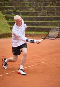 Idrottare i vit långärmad tröja med svarta detaljer, svarta shorts och röd/svarta tennisskor, som utför en forehand-slag på en grusplan.