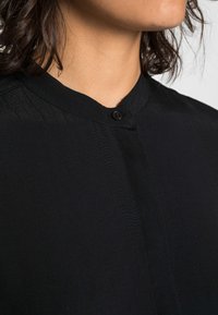 Camisa preta com colarinho tipo camisola, fecho de um único botão preto e um tecido liso e texturizado com um leve brilho.