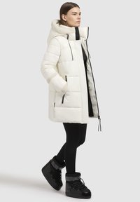 Vit pufferjacka med foder av konstpäls i huvan och ärmsluten, med dragkedjefickor och längd till mitten av låret. Svarta stövlar kompletterar outfiten.