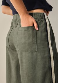Green linen pants with side pockets, elastic waistband, and contrasting side stripe. Fabric texture appears lightweight and breathable.