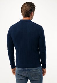 Námořnický svetr z ribrového úpletu s límcem, dlouhými rukávy a přiléhavým pasem. Nosený s světle modrými džíny. Žádné viditelné logo ani vzory.
