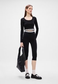 Μαύρο μπλούζα μακρυμάνικη και κοντές ποδηλατικές βερμούδες με λευκή λογότυπη ζώνη. Το μοντέλο κρατάει μια μαύρη τσάντα, φορώντας λευκές κάλτσες και μαύρα παπούτσια.