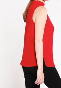 Top rosso senza maniche in tessuto leggero, caratterizzato da un collo alto e spacchi laterali; texture liscia con una vestibilità rilassata.