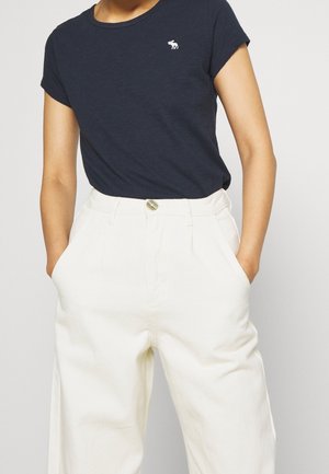 Chemise bleu marine à manches courtes avec logo élan blanc, rentrée dans un pantalon blanc taille haute, les mains dans les poches.