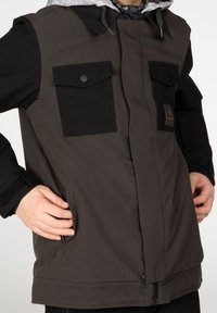 Giacca a zip marrone e nera con cappuccio grigio, due tasche sul petto e tasche laterali; presenta un tessuto testurizzato con cuciture visibili.