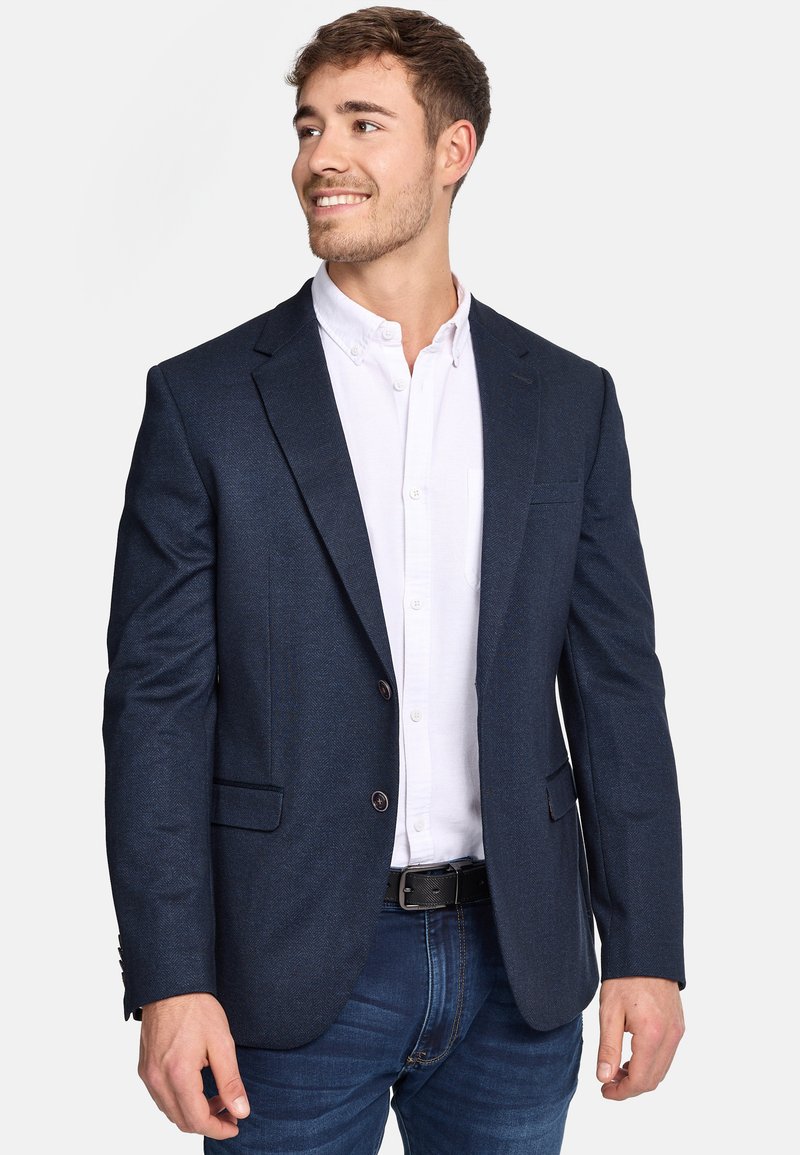 Marineblauer Blazer mit strukturiertem Muster, mit Revers, zwei Knöpfen und Seitentaschen, getragen über einem weißen Hemd mit Knopf und Jeans.