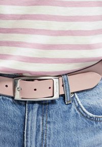 Cinturón de cuero rosa claro con una hebilla rectangular plateada, abrochado a unos vaqueros de mezclilla con un acabado azul desgastado y costuras amarillas.