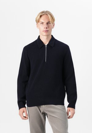 Navy knitted pullover with a ribbed texture, featuring a collar and a short front zipper, paired with light-colored trousers.
