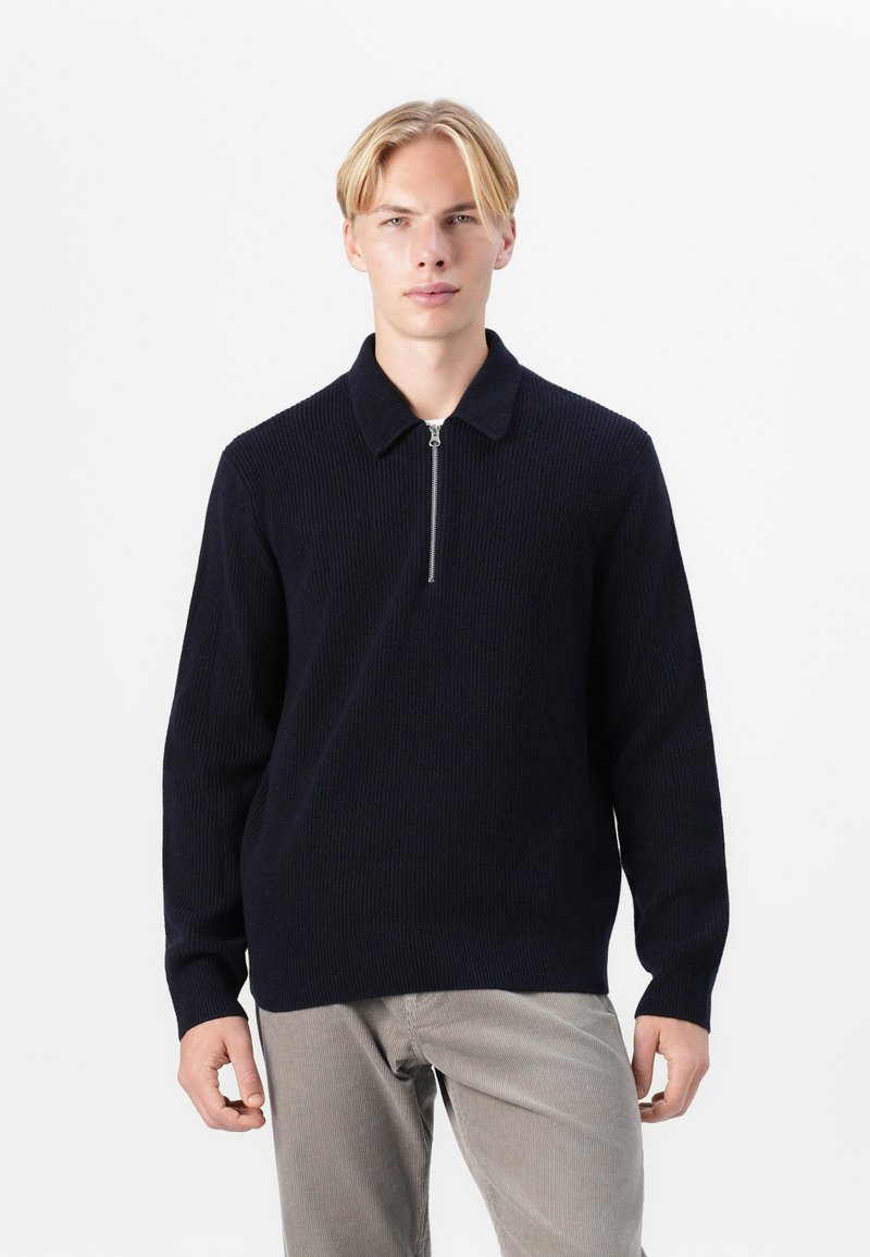 Tumedast sinise värvi kudumipullover ribitud tekstuuriga, millel on kaelus ja lühike esine tõmblukk, paired heledate pükste jaoks.