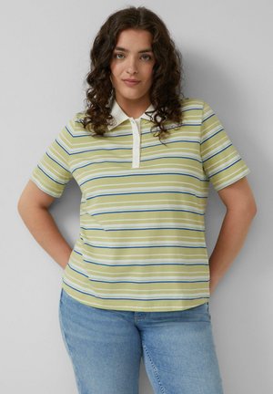 Junge Frau mit lockigem Haar, die ein grün-weiß-blau gestreiftes Poloshirt und helle blaue Jeans trägt, steht mit den Händen hinter dem Rücken.