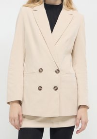 Beżowa kurtka ze sztruksu o fasonie na dwa rzędy guzików, z dwoma brązowymi guzikami i bocznymi kieszeniami. Teksturowana tkanina i dopasowany krój. Długie rękawy.