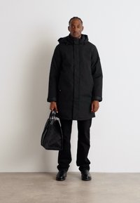 Schwarzer, isolierter langer Mantel mit Kapuze, getragen über schwarzen Hosen und Schuhen. Die Person hält eine strukturierte schwarze Tasche und steht vor einem neutralen Hintergrund.