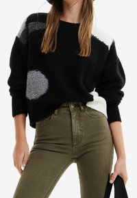 Czarny sweter z szaro-białymi geometrycznymi wzorami, ściągacze na rękawach, w zestawieniu z oliwkowo zielonymi jeansami z wysokim stanem. Miękka faktura i luźny krój.
