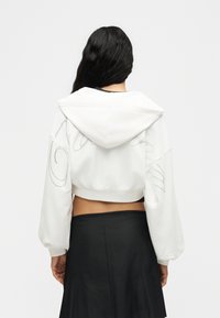 Γυναίκα με μακριά σκούρα μαλλιά που φοράει λευκό cropped φούτερ με λεπτομέρειες διάτρητου κειμένου και μαύρη ψηλόμεση φούστα, εμφανίζεται από πίσω.