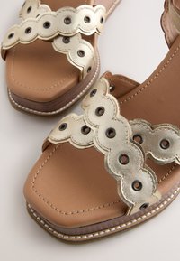Sandales en cuir beige avec des brides métalliques festonnées comprenant des œillets noirs et des coutures blanches visibles le long des bords de la semelle.