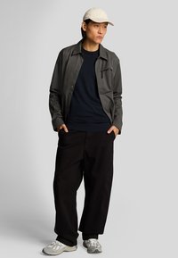 Giacca grigia abbottonata sopra un maglione blu navy; pantaloni neri larghi; cappellino beige; scarpe grigie e bianche con suole testurizzate.