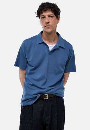 Homme aux cheveux bouclés et foncés portant un polo bleu à manches courtes par-dessus un sous-vêtement blanc et un pantalon sombre, debout devant un fond uni.