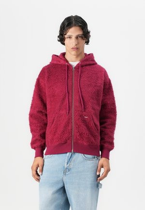 Red, fuzzy zip-up hoodie with drawstrings, round pockets, ribbed cuffs, and hem. Paired with light blue jeans.