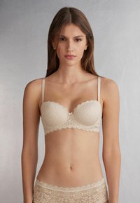 Beige kanten bh met geborduurde randen en een bloemmotief, voorzien van verstelbare bandjes en een klein hartaccent in het midden.