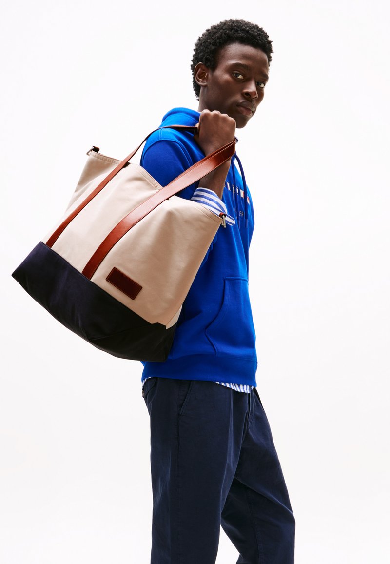Sac fourre-tout beige avec un fond bleu foncé et des poignées en cuir marron. Présente une texture lisse et un design minimaliste. Polyvalent et spacieux.