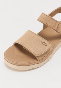 Tan suede-sandaali, jossa on kaksi säädettävää hihnaa ja pehmustettu pohja. Varustettu kevyellä beigeillä kumipohjalla ja painettulla logo-yksityiskohdalla.