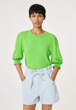 Vrouw draagt een felgroene top met driekwart mouwen en gouden knopen en lichtblauwe high waisted paperbag-short met een gestrikte riem.