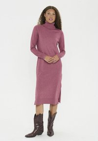 Roosa pehme kudumisega turtleneck kleit, millel on keskmine pikkus ja külgmised lõhikud, koos pruunide kauboi saapatega. Kanga pehmus ja soonilised varrukad toovad esile elegantse stiili.