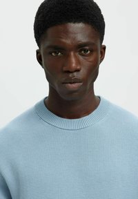 Svijetloplavi pleteni pulover s okruglim izrezom; sadrži teksturirani uzorak i rebraste rukave. Silueta je uska, a tkanina izgleda mekano.