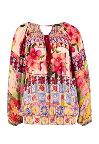 Blouse style bohème à manches longues, motifs floraux et géométriques roses, encolure froncée avec liens à pompons rouges, et ourlet et poignets élastiques.