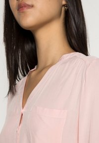 Blusa rosa chiaro con scollatura a V, texture morbida, chiusura a bottoni e una tasca singola sul lato sinistro. Presenta una vestibilità rilassata e cuciture delicate.