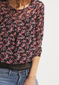 Blouse noire à fleurs en tissu léger et transparent, avec des motifs floraux roses et turquoise et des manches trois-quarts. Coupe ample.