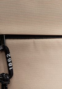 Beiger Canvas-Stoff mit strukturierten Oberflächen, ausgestattet mit einem schwarzen Reißverschluss und Hardware, die die Zahl "1862" zeigt, befestigt an einem schwarzen Band.