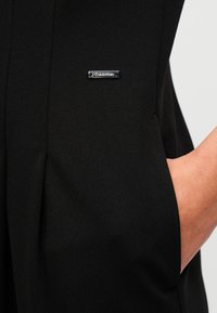 Μαύρο ύφασμα με λεία υφή που διαθέτει μεταλλικό λογότυπο Calvin Klein, καθώς και λεπτομέρεια με τσέπη. Ο σχεδιασμός είναι μινιμαλιστικός και κομψός.