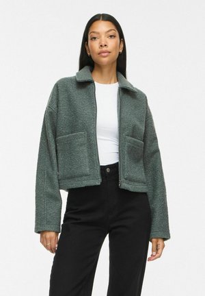 Femme aux longs cheveux foncés portant une veste polaire grise zippée, une chemise blanche et un pantalon noir taille haute sur un fond uni.