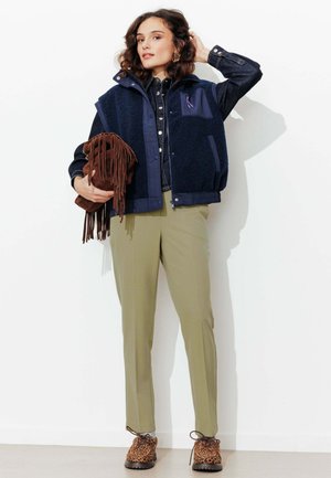 Femme portant un gilet en polaire bleu marine et une veste en jean, tenant un sac marron à franges, avec un pantalon olive et des chaussures imprimé léopard, debout contre un mur blanc.