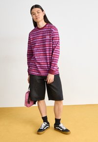 Cămașă cu mânecă lungă în dungi magenta, cu guler rotund, asortată cu șorturi negre din denim și adidași negri, ținând o geantă roz. Fundal galben.