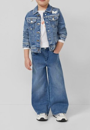 Kind draagt een blauwe spijkerjack met vlinderapplicatie, wit shirt, brede blauwe jeans en witte sneakers, staat met één hand in de zak.
