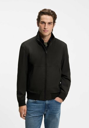 Ung mand iført en sort lynlåstrøje over en skjorte og blå jeans, stående med den ene hånd i lommen mod en hvid baggrund.