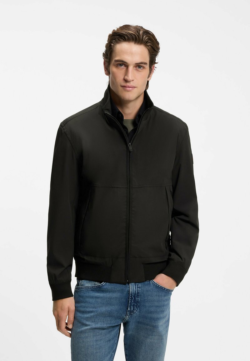 Jeune homme portant une veste noire zippée par-dessus une chemise et un jean bleu, debout avec une main dans la poche sur fond blanc.